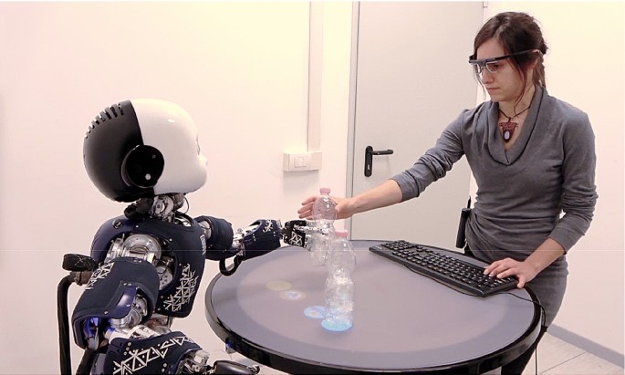 A humanoid robot with a baby-like face interacts with a woman at a table during an AI experiment