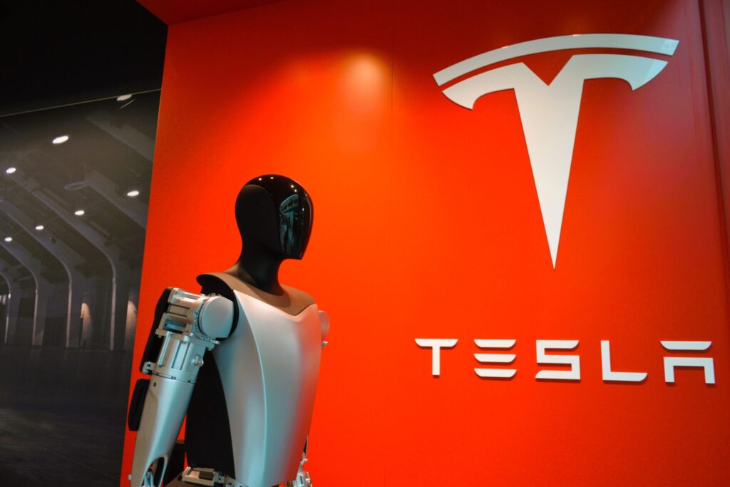 Humanoid Optimus robot standing in front of Tesla logo showcasing futuristic robotic technology.
