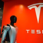 Humanoid Optimus robot standing in front of Tesla logo showcasing futuristic robotic technology.