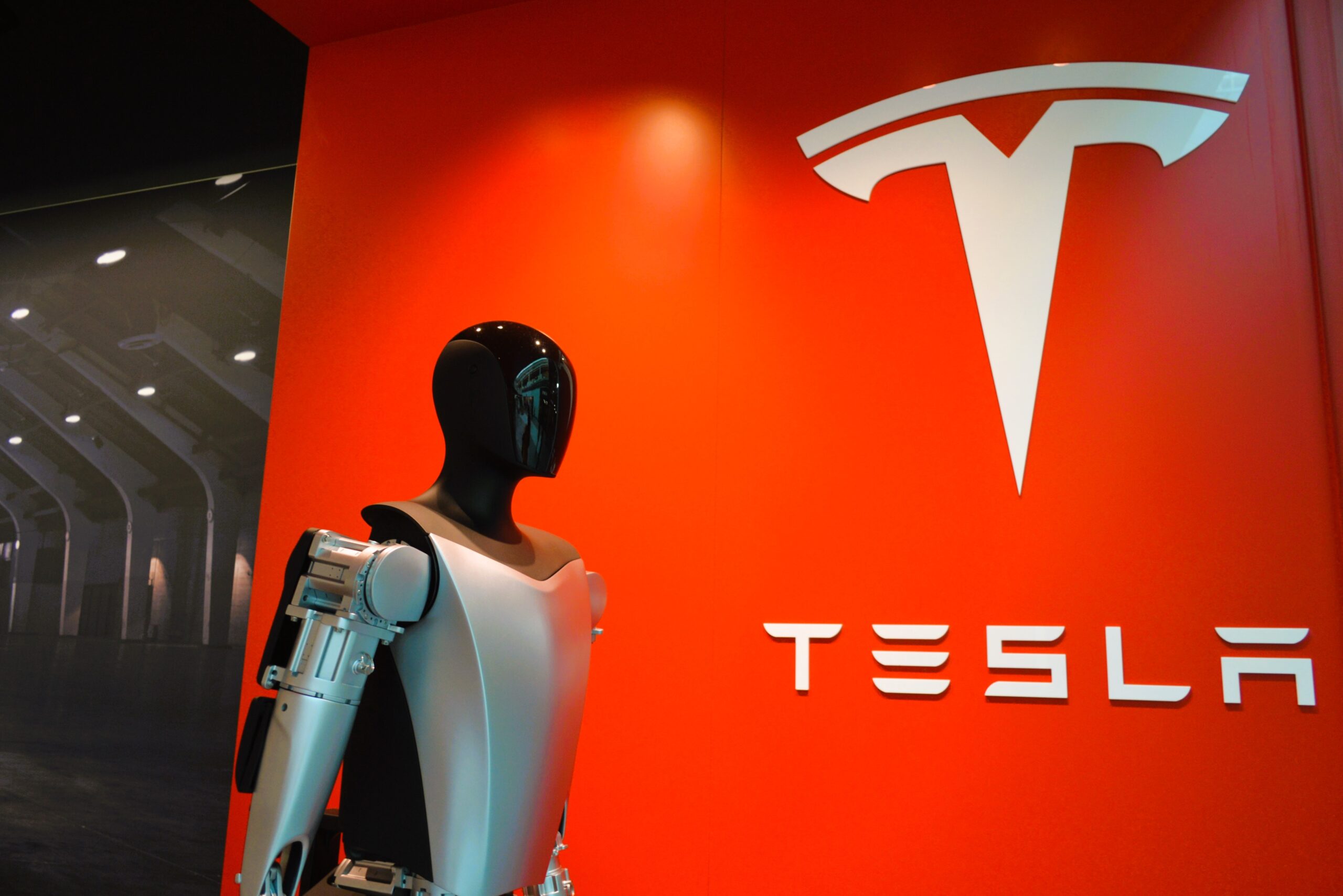 Humanoid Optimus robot standing in front of Tesla logo showcasing futuristic robotic technology.