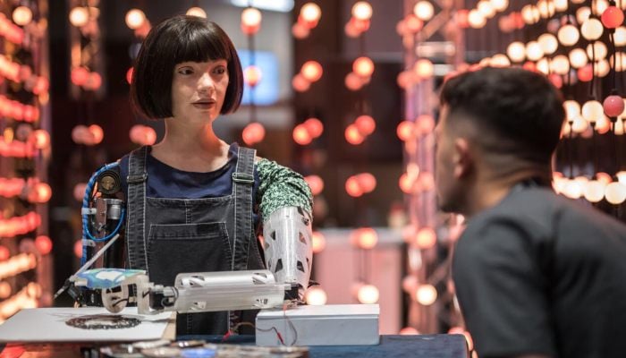 Humanoid robot artist Ai-Da interacting with a visitor while painting at an exhibition, surrounded by lights