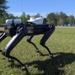 Asylon's robot guard dog standing on grassy field during daytime, equipped with surveillance hardware and marked with security warnings.