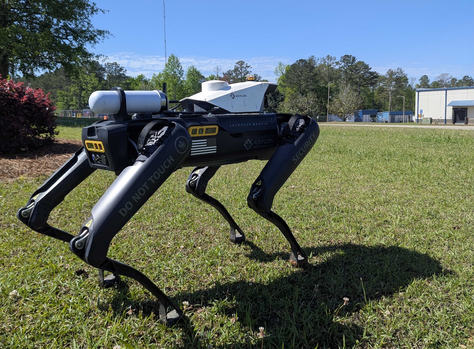 Asylon's robot guard dog standing on grassy field during daytime, equipped with surveillance hardware and marked with security warnings.