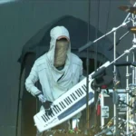 Funky humanoid robot rocks the keys during a live music festival performance, wearing a hoodie and cap.