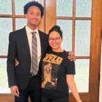 A young man wearing a black suit and striped tie stands beside a smiling woman in a graphic T-shirt as they pose together in front of wooden glass-paneled doors indoors.