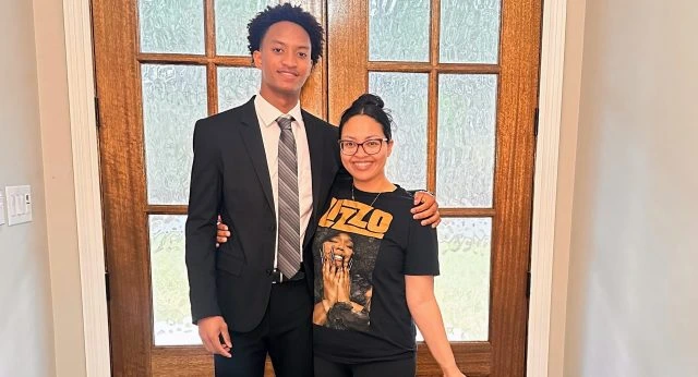 A young man wearing a black suit and striped tie stands beside a smiling woman in a graphic T-shirt as they pose together in front of wooden glass-paneled doors indoors.