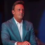 Karl Ravech appears on an ESPN broadcast set wearing a light blue blazer and headset during a live sports television production.