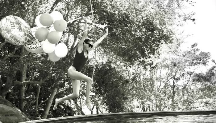 Meghan Markle jumps into a swimming pool holding birthday balloons while celebrating her 45th birthday in a black-and-white photo.