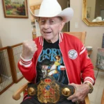 Dory Funk Jr. wearing a white cowboy hat and red jacket while holding the NWA World Heavyweight Championship belt and raising his fist in a recent photo.