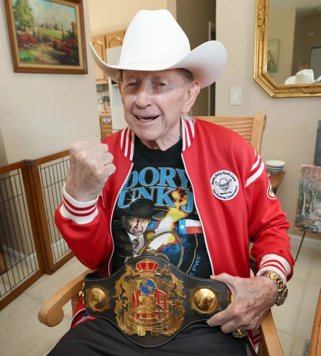 Dory Funk Jr. wearing a white cowboy hat and red jacket while holding the NWA World Heavyweight Championship belt and raising his fist in a recent photo.